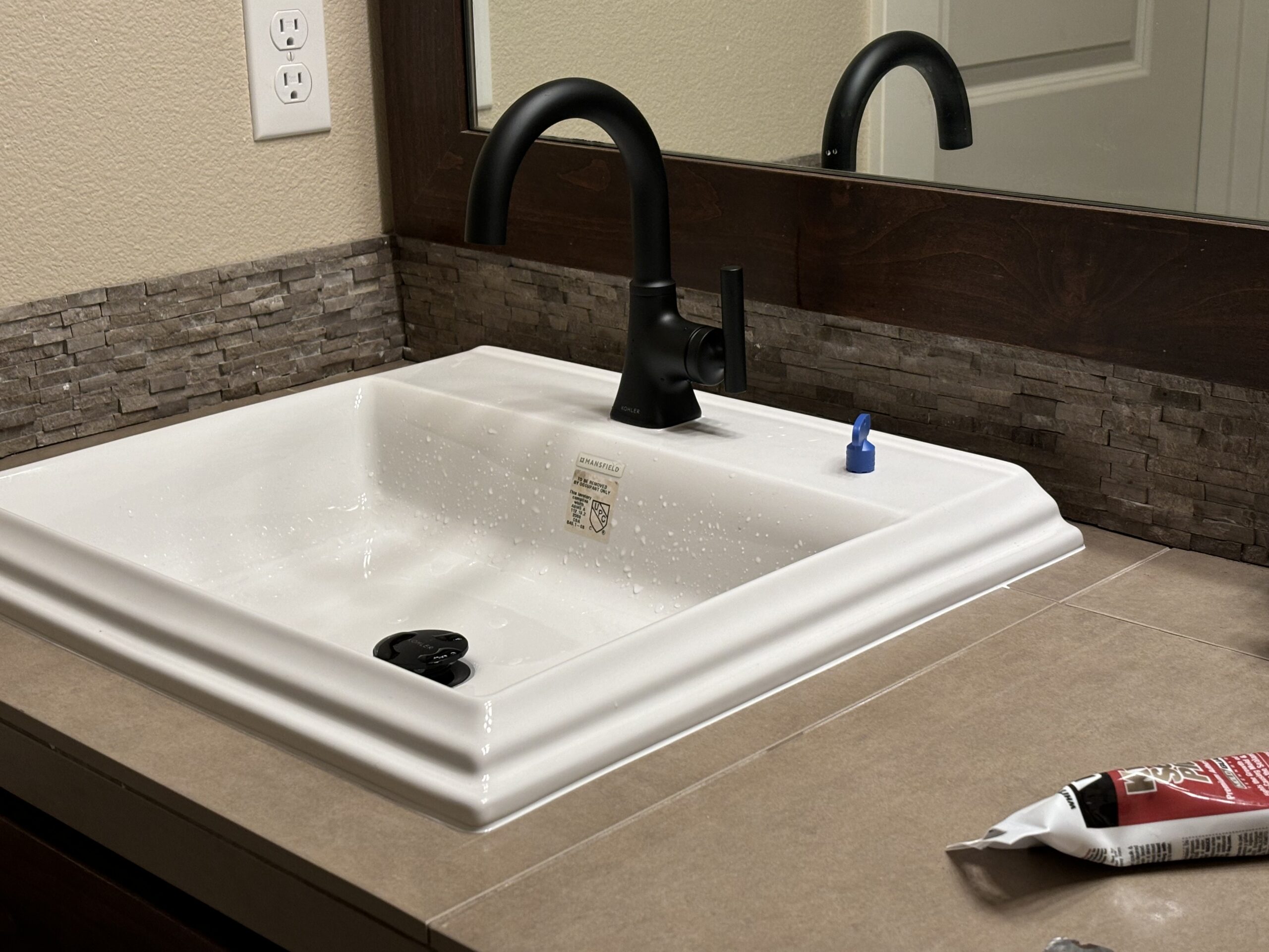 A white rectangular sink with a black faucet is installed on a tan countertop, with a mirror and stone tile backsplash behind it. A blue cap and a tube of sealant are on the counter.