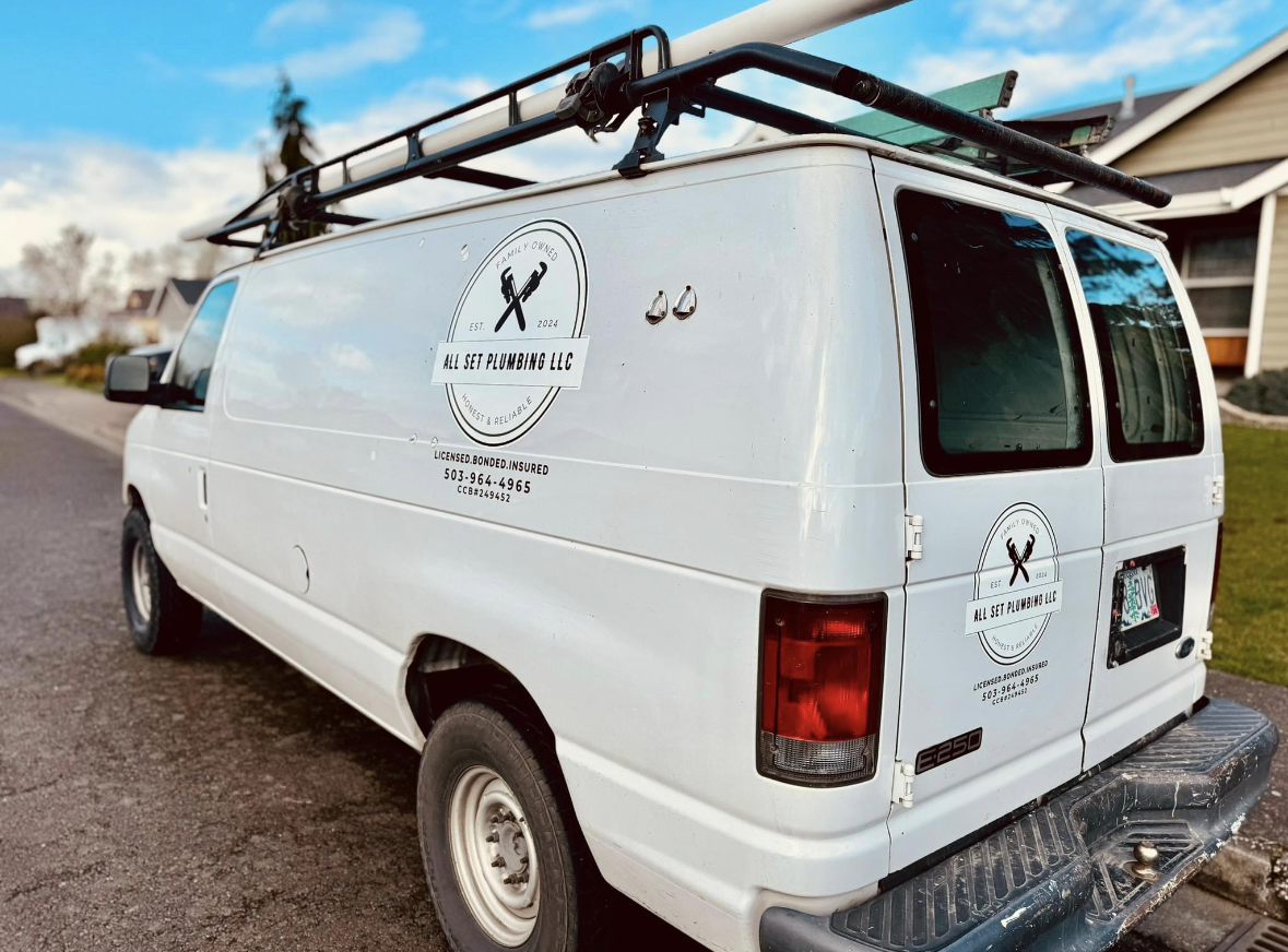 A white plumbing service van with company logos on the side and rear is parked on a residential street. A ladder rack is mounted on the roof.