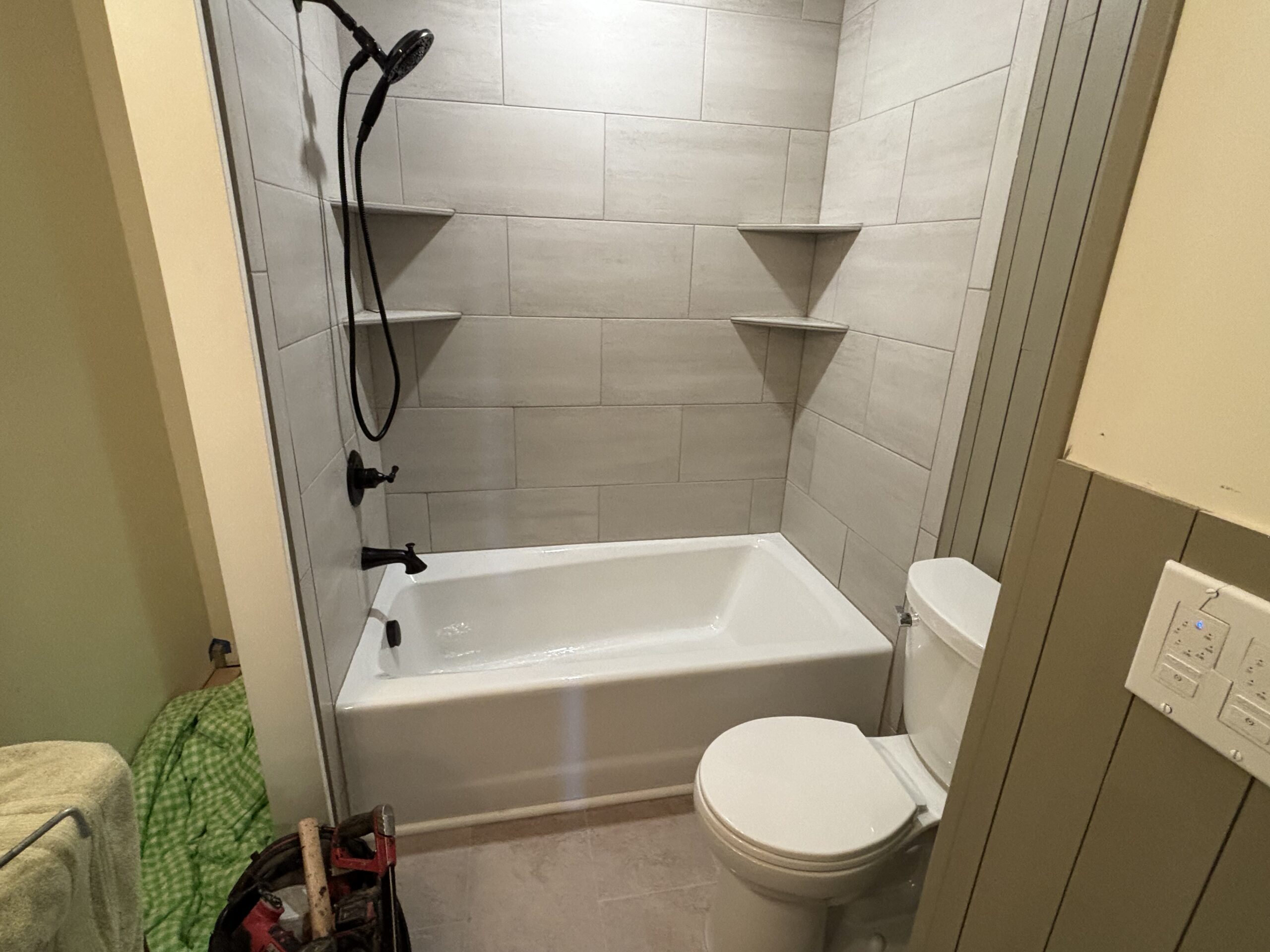 A bathroom with a white bathtub, gray tiled walls, three built-in corner shelves, a black showerhead, and a white toilet next to a light switch panel.