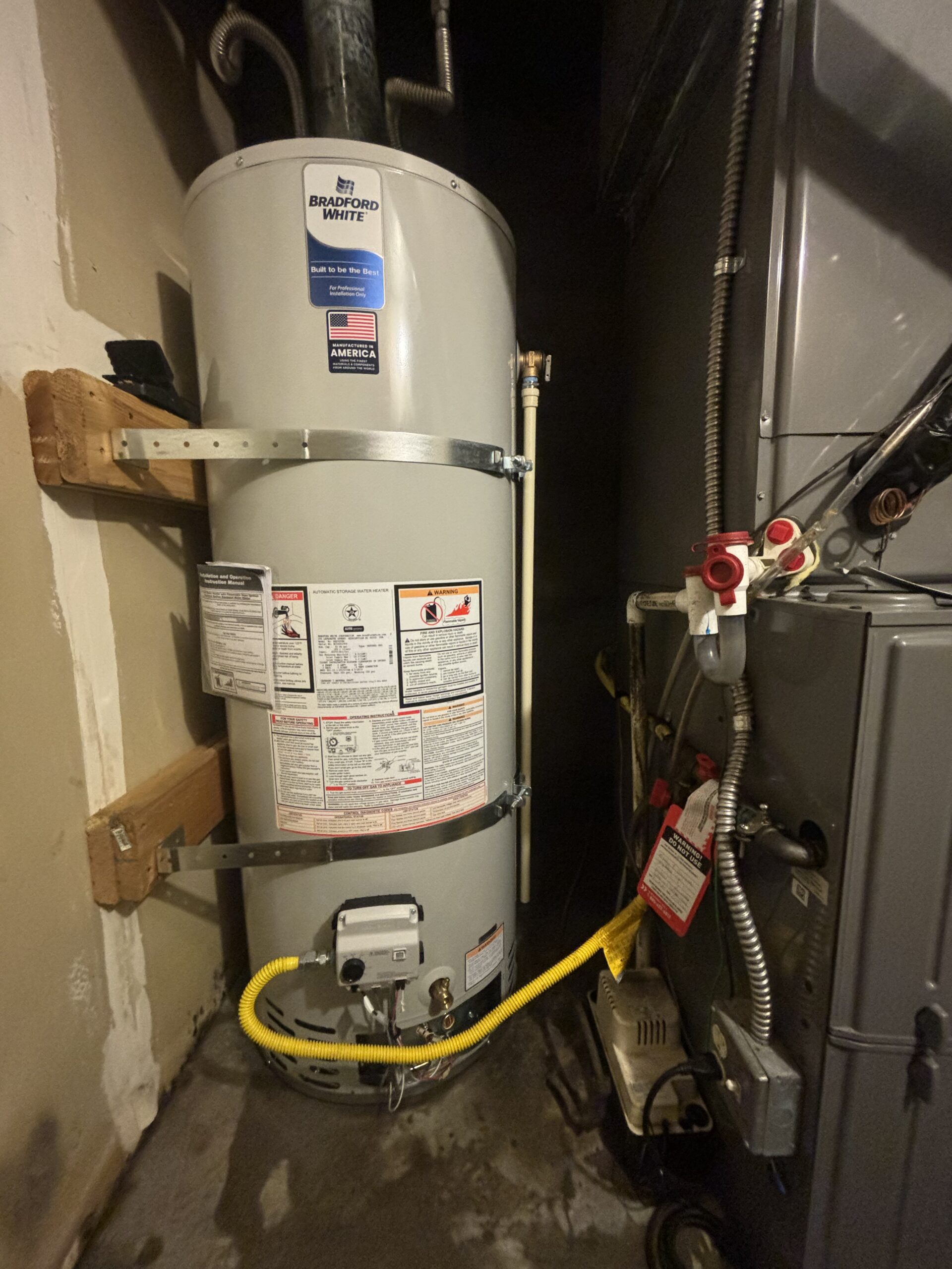 A residential water heater installed next to a furnace in a utility closet, with visible warning labels, pipes, and wiring.