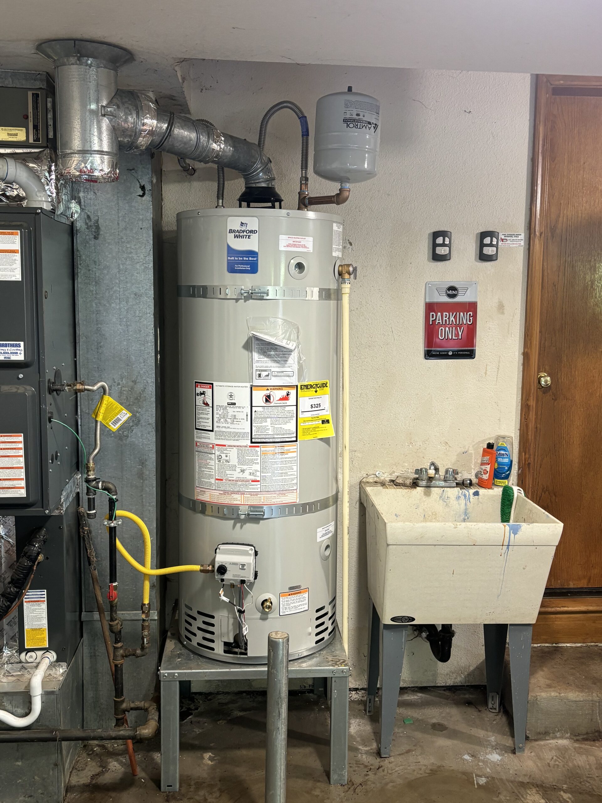 A gas water heater next to a utility sink in a basement, with pipes, warning labels, meters, and a Parking Only sign on the wall.