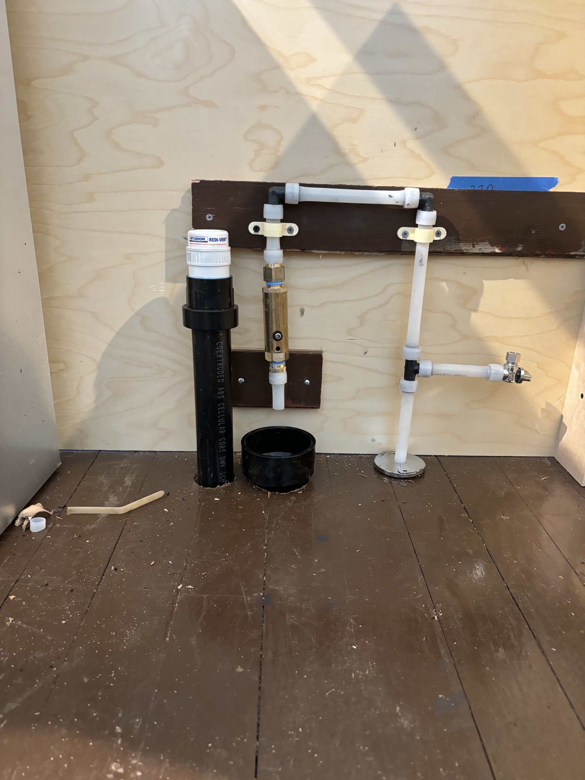 Plumbing pipes and fixtures mounted on a plywood wall with a black drain pipe, a white valve, and a water meter above a wooden floor.